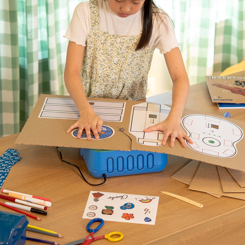 A child wearing a floral dress sits behind a wooden desk using the ChompSaw to cut out a cardboard ukulele from the Inventor's Club. She's surrounded by markers, scissors, and other school supplies in a brightly lit room.