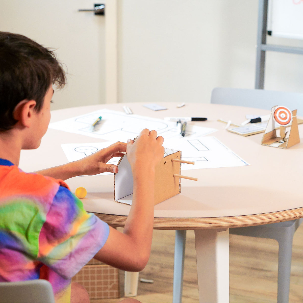 A child in a colorful tie-dye shirt sits at a light wood table, building a small cardboard catapult with a spinning target from Chompshop's Inventor's Club, Papers and small craft items are scattered on the table in a bright indoor room.