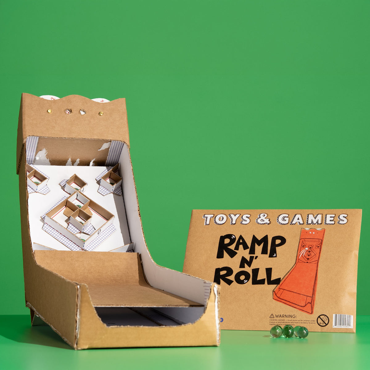 A cardboard Ramp 'N Roll toy game setup on a bright green background, with the open cardboard machine showing printed track pieces inside and a branded cardboard box beside it with three green marbles.