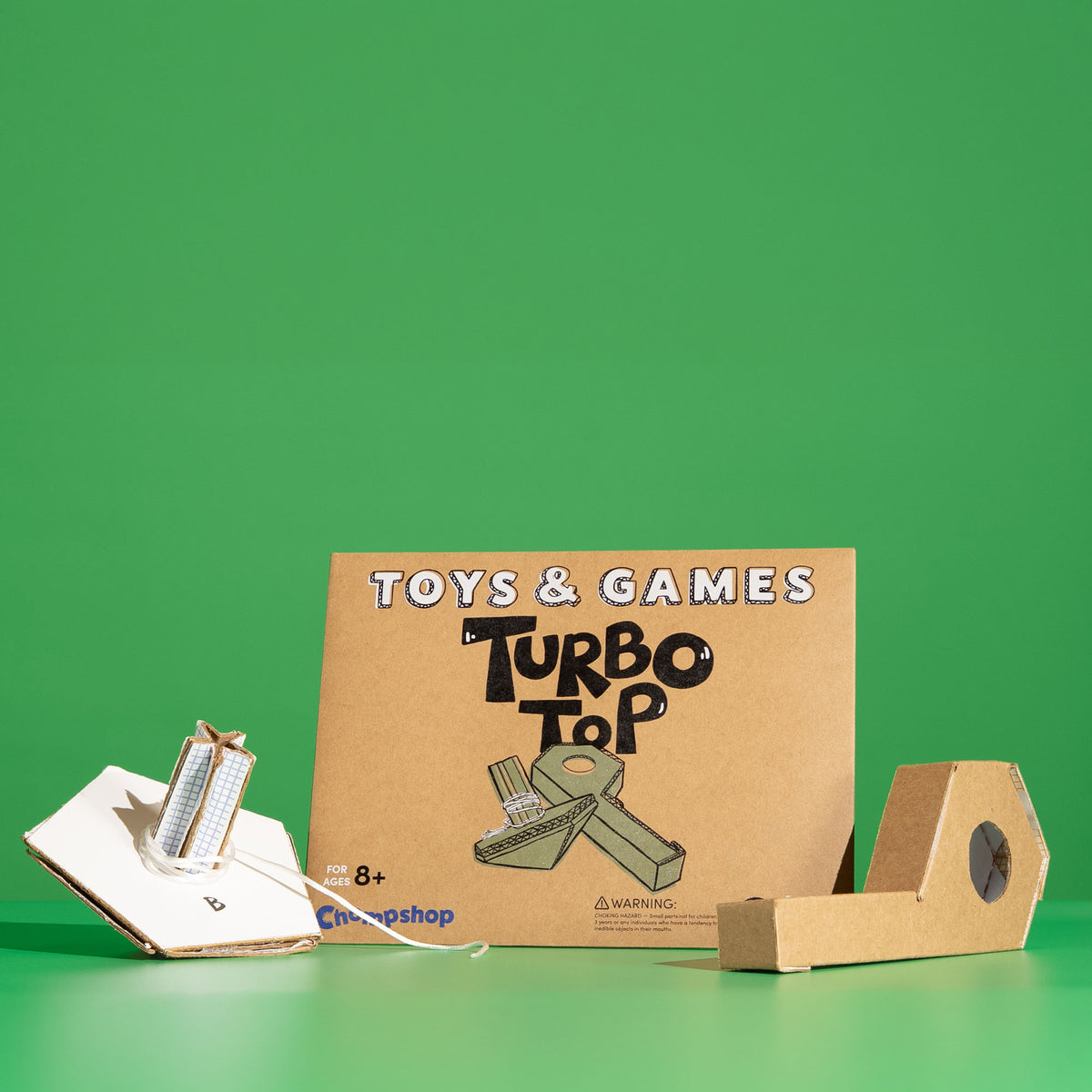 A cardboard toy kit labeled 'TOYS & GAMES Turbo Top' stands on a green surface against a solid green background.