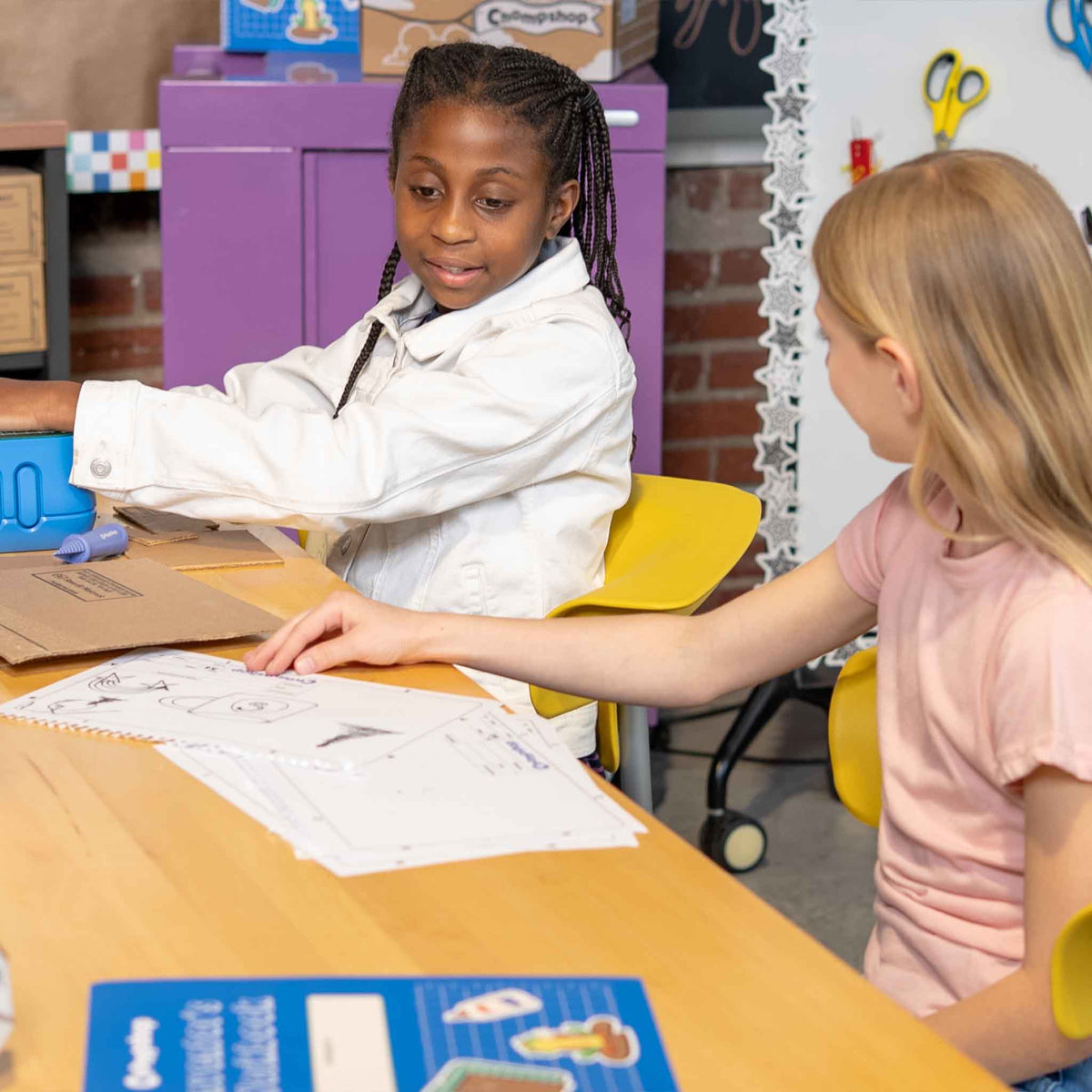 Two children sitting at a table in a classroom setting, engaged in an activity.