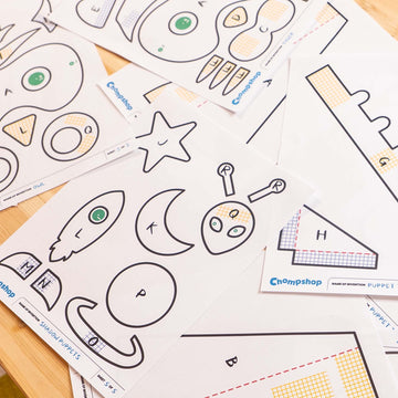 Several printed paper templates for assembling a shadow puppet show the cut-out parts on a light wood surface. The sheets contain simple black outline shapes and some colored sections, with the brand name Chompshop visible on the sheets.