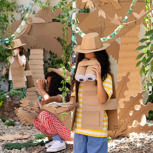 Children outdoors crafting with cardboard, wearing cardboard safari hats and vests, with one child holding cardboard binoculars in a garden setting.