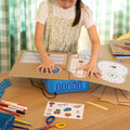 A child wearing a floral dress sits behind a wooden desk using the ChompSaw to cut out a cardboard ukulele from the Inventor's Club. She's surrounded by markers, scissors, and other school supplies in a brightly lit room.