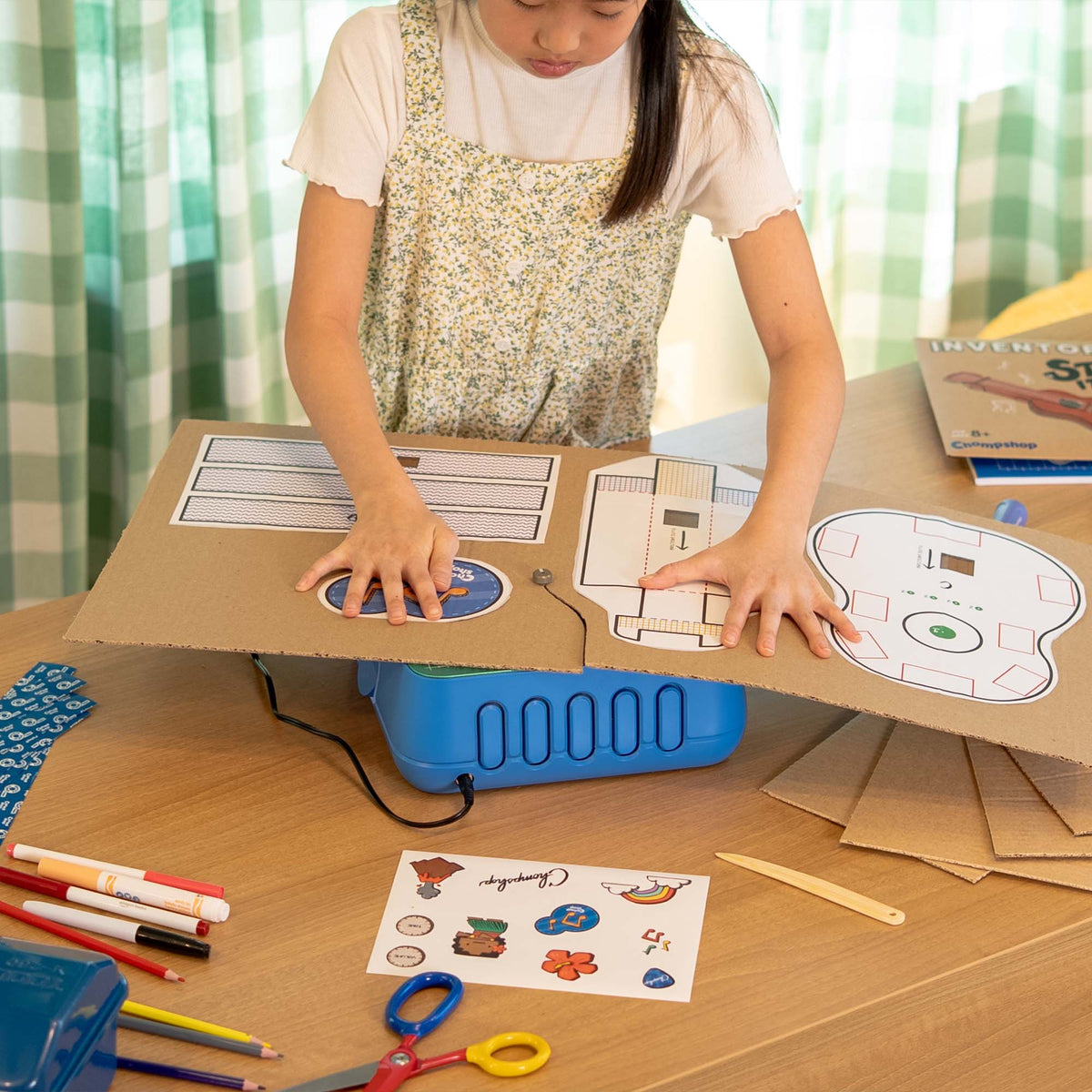 A child wearing a floral dress sits behind a wooden desk using the ChompSaw to cut out a cardboard ukulele from the Inventor's Club. She's surrounded by markers, scissors, and other school supplies in a brightly lit room.