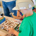 A child wearing a cardboard hat and green shirt plays with a small cardboard maze on a table in a workshop setting.