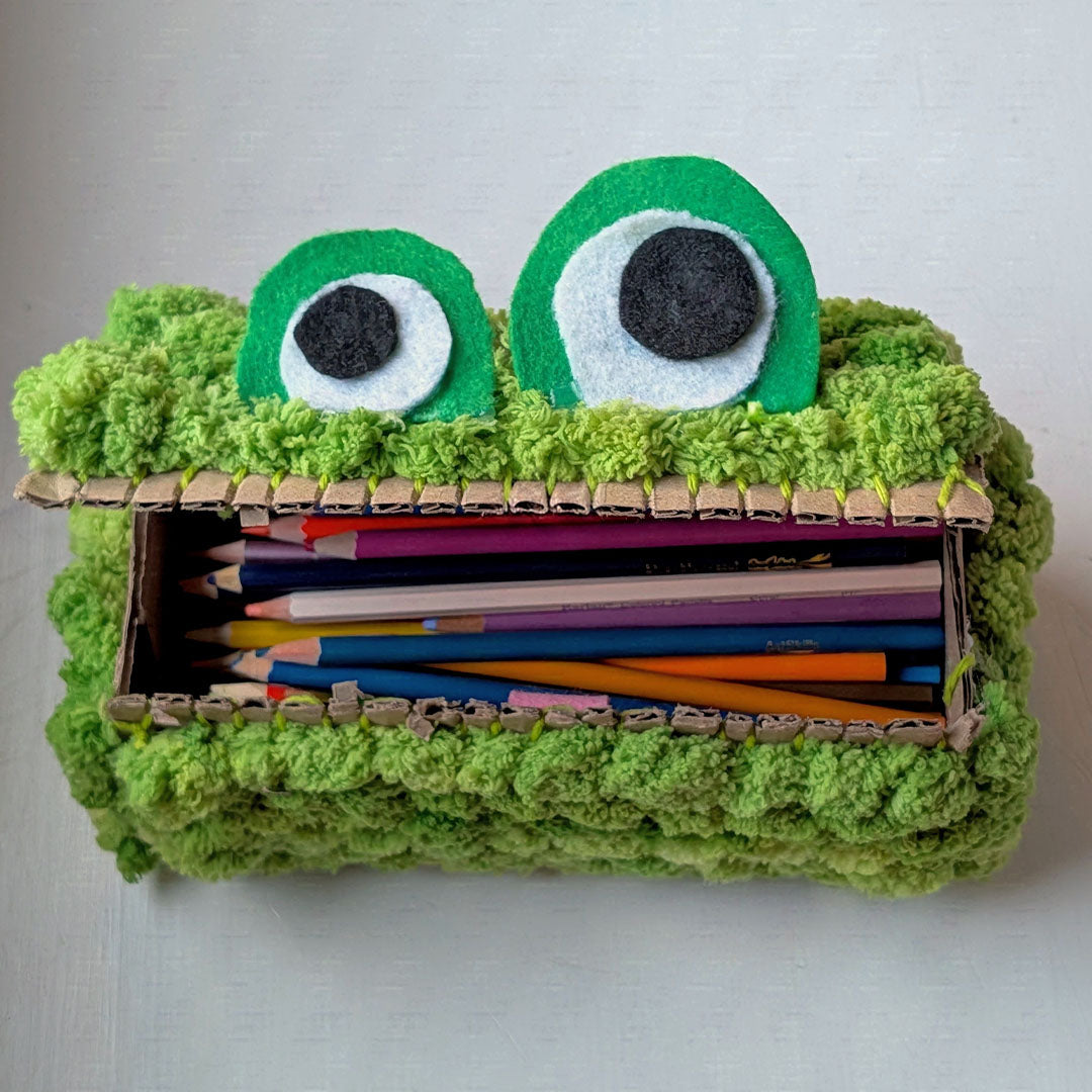 A handmade monster-themed pencil case made from a cardboard box, covered in textured green yarn, with two large felt eyes on top, shown open with colored pencils inside on a light background.