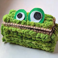 A handmade green monster face tissue box cover made from textured fuzzy yarn, featuring two large felt eyes with black and white centers and a cardboard mouth.