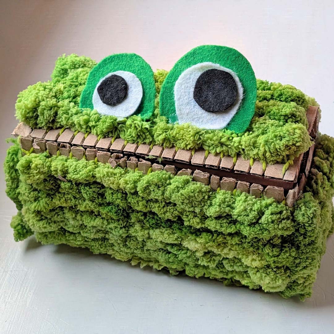 A handmade green monster face tissue box cover made from textured fuzzy yarn, featuring two large felt eyes with black and white centers and a cardboard mouth.