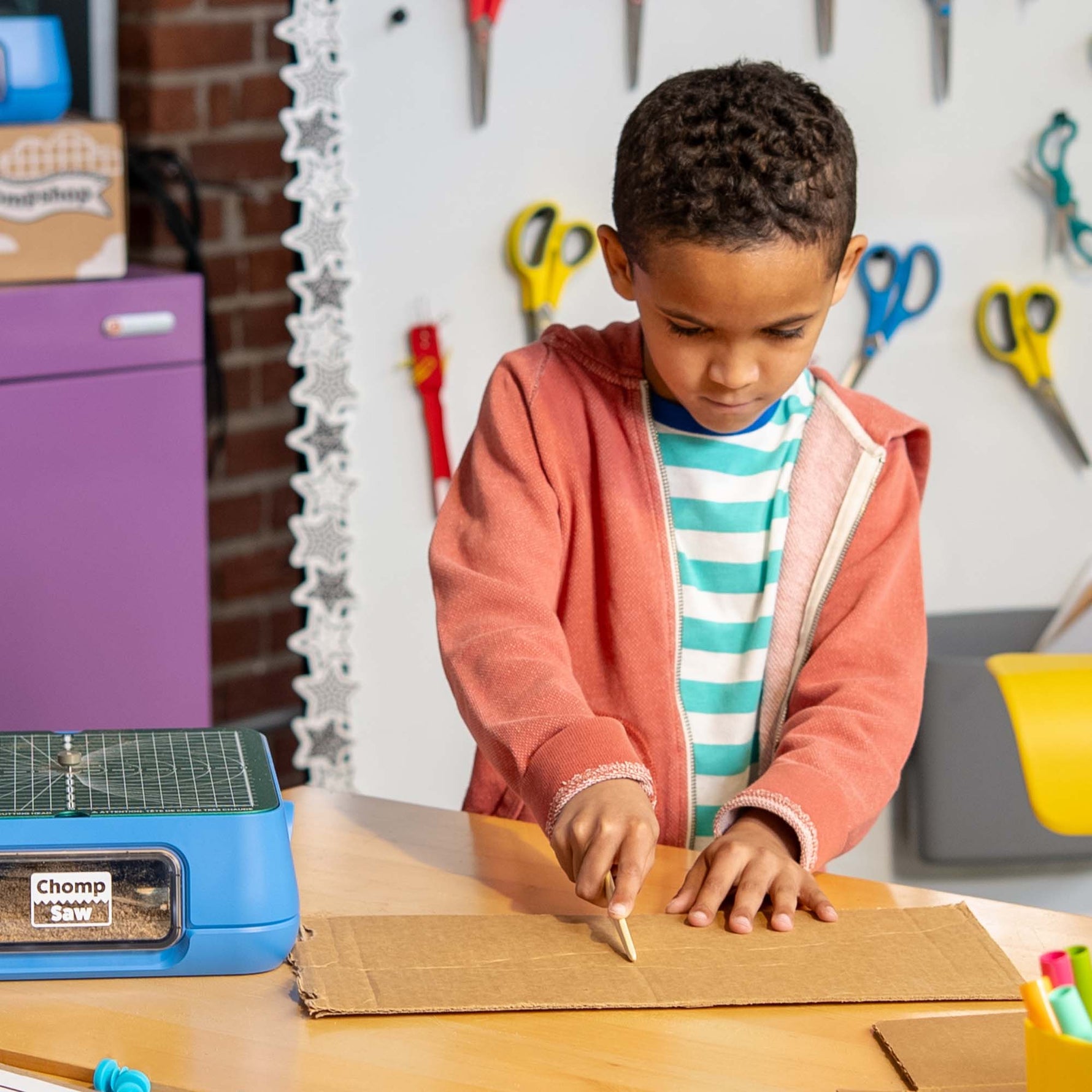 Child using Chompshop's Scoring Tool accessory for folding and creasing cardboard in a classroom.