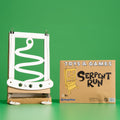 A cardboard toy set called 'Serpent Run' by Chompshop, featuring a white snake-shaped track on a brown cardboard base, shown next to its brown product box against a solid green background.