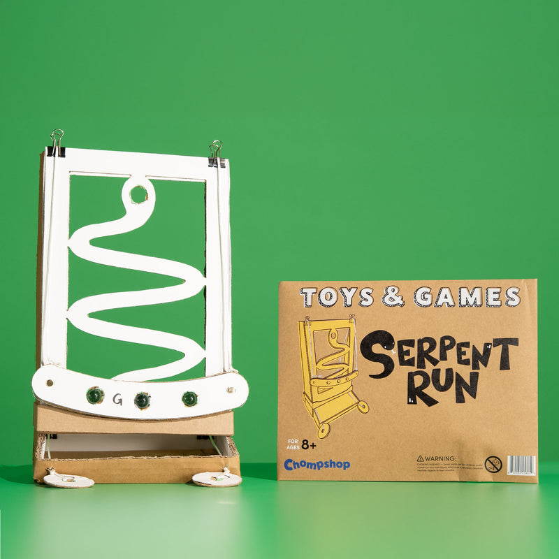 A cardboard toy set called 'Serpent Run' by Chompshop, featuring a white snake-shaped track on a brown cardboard base, shown next to its brown product box against a solid green background.