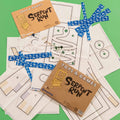 Toy and game set laid out on a bright green background, including printed patterns, a cardboard package labeled 'Toys & Games Serpent Run,' blue patterned tape strips, white string, and marbles. The visible brand on the packaging is CircusShop.