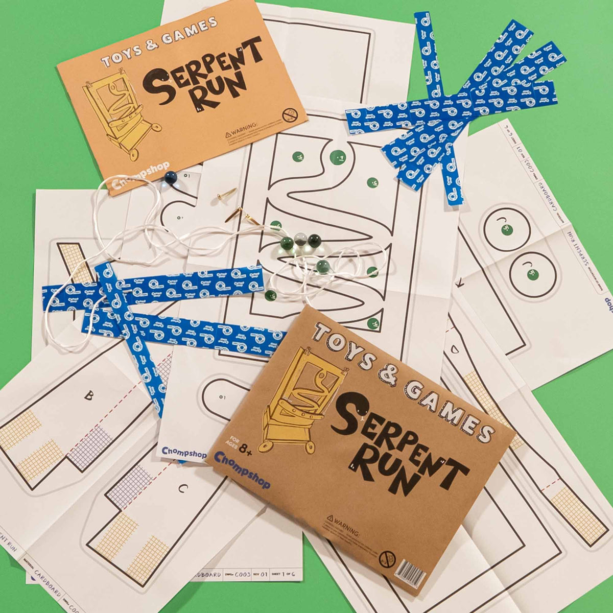 Toy and game set laid out on a bright green background, including printed patterns, a cardboard package labeled 'Toys & Games Serpent Run,' blue patterned tape strips, white string, and marbles. The visible brand on the packaging is CircusShop.