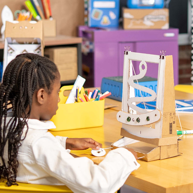A child in a white shirt sits at a yellow desk in a classroom, interacting with a cardboard snake game. The background shows colorful storage bins and school supplies.