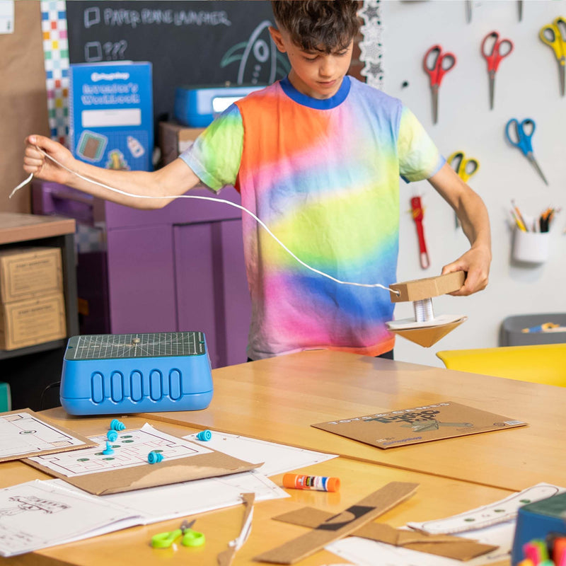 A boy in a rainbow tie-dye t-shirt is building a cardboard spinning top in a classroom setting, holding a white string and a cardboard structure. The desk in front of him is cluttered with craft supplies, including a blue plastic mold, papers, and scissors.