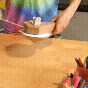 A handmade cardboard turbo top spinning on a white plate on a wooden desk.
