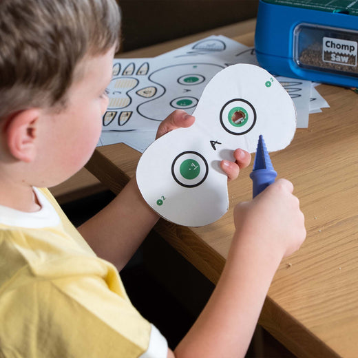 A child in a yellow shirt is shown from the side, holding a cardboard pattern and using a Chompshop hole punch tool. 