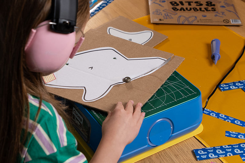 A child wearing pink earmuffs and a striped shirt uses the ChompSaw to cut an Explorer's Club project template shaped like a shark. The workspace is covered blue strips of tape, and a Chompshop hole punch tool, with a cardboard package labeled 'BITS & BAUBLES' in the background.